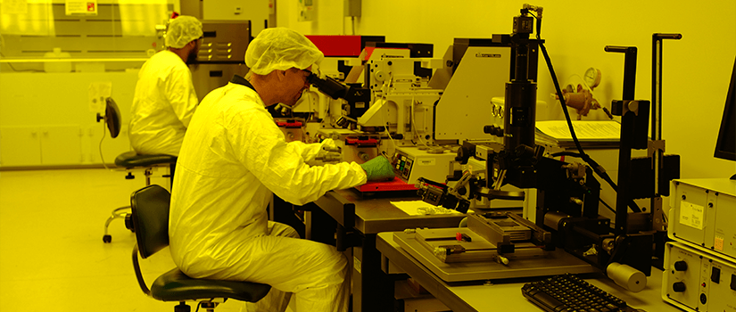 Two researchers in white lab coats looking through microscopes in a yellow-colored room for chip packaging.