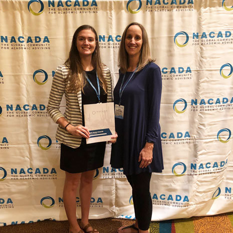 Rachel is pictured wearing a black and white striped dress and holding a copy of the NACADA certificate of merit. Jane is to Rachel's left and is wearing a long blue shirt and black leggings. Both women are smiling. 