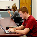 A male student sits at a laptop computer. He is wearing a garnet tshirt and is looking at his computer screen.