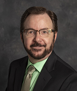 bill is pictured from the waist up wearing a black jacket, green shirt, and tie. He has dark brown hair, glasses, and is smiling. 