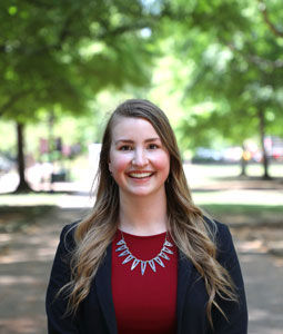 Alexa is pictured outside with green trees blurred in the background. She is wearing a garnet shirt and black blazer with a silver necklace. Her long blonde/brown hair is curled and she is smiling. 