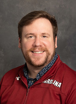 Cam is pictured from the shoulders up wearing a garnet pullover with Carolina written on the shoulder. He has pale skin, brown hair, and is smiling. 