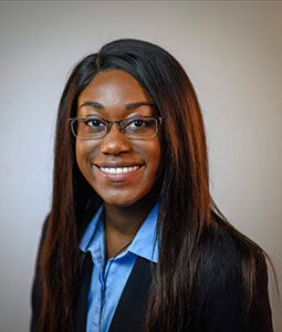 Kiisha is pictured from the shoulders up wearing a blue shirt and black jacket. She has brown skin, long hair, is wearing glasses, and is smiling 