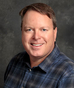 Keith is pictured from the shoulders up in a blue plaid shirt. He has short brown hair and is smiling. 