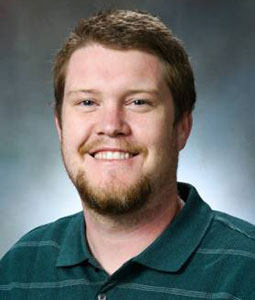 Chris is picture from the shoulders up wearing a green collared shirt. He has blondish-brown hair and a beard of the same color and smiling. 