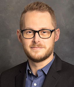 Ben is pictured from the shoulders up wearing a blue shirt and black blazer. Ben has fair skin, blond hair, a blonde beard and is wearing glasses. 