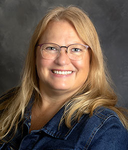 Mary Jo is picture from the shoulders up. She has long blonde hair, is wearing glasses and is smiling. 