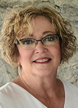 Susan is picture from the shoulders up. She has curly, blonde hair and is wearing a white shirt and glasses. She is smiling. 