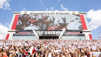 The Williams-Brice Jumbotron showing the team preparing to take the field, with a full student section cheering loudly with tree and gates mark.