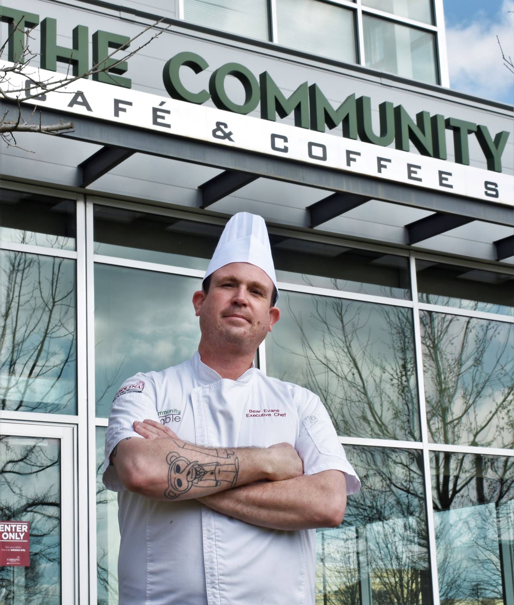 Chef Bear Evans standing with his arms crossed outside in from of a sign that reads "The Community Table Cafe and Coffee Shop"