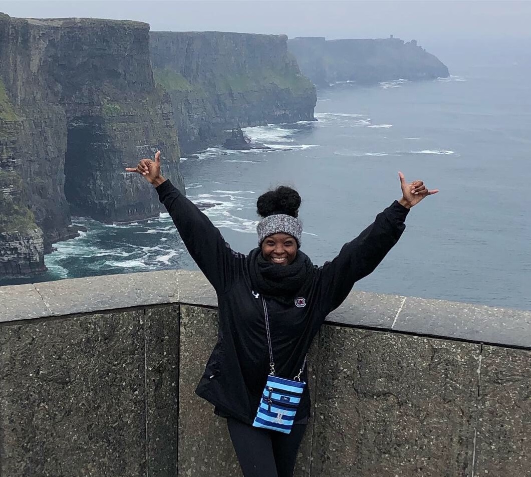 Student poses in front of a body of water and cliffs