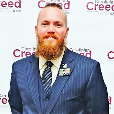 A man with a red beard is standing in front of a Carolinian Creed banner in a professional suit.