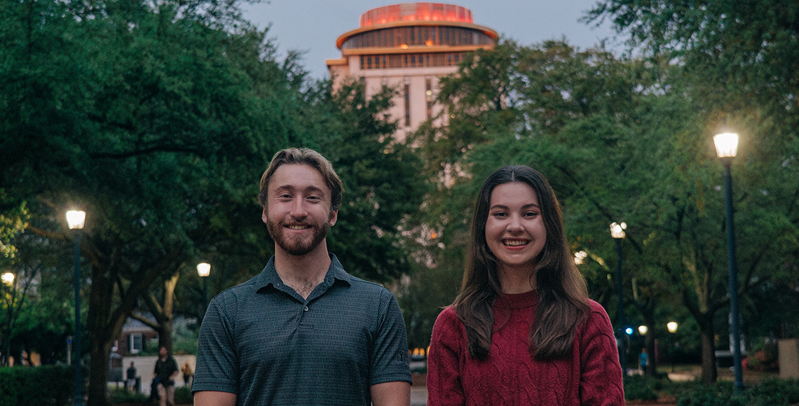 Two students standing in front of a tall building with a garnet light behind them.