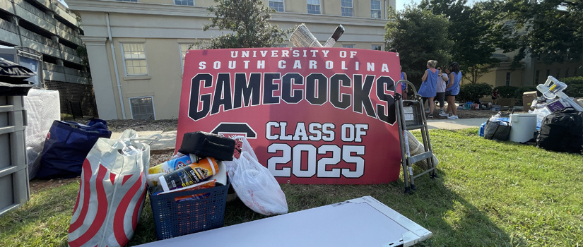 A stack of student room supplies sit outside of a four story high residence hall with a Gamecocks class of 2025 sign