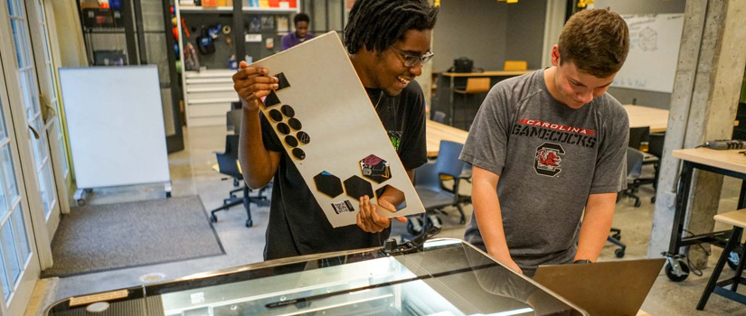 Two students hold boards as they load them into a cutting printer in the Rhodos Fellows Makers Space.