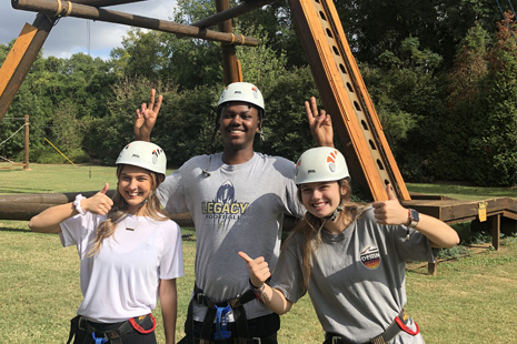 Three students give a thumbs up wearing helments in front of the ropes course at UofSC