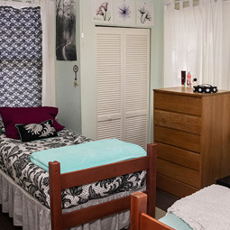 A fully furnished bedroom. To the left is a bed with a black and white patterned bedspread and a blue blanket on top. In the middle of the room is a dresser and closet. Two windows in the room are covered with curtains. 