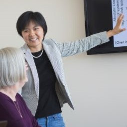 Instructor smiling, standing up in front of a class teaching and pointing to a whiteboard. 