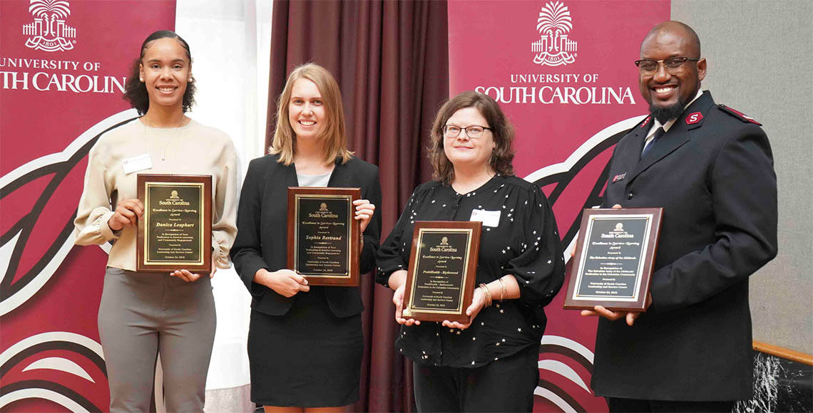 Four people hold award plaques and smile in front of University of South Carolina banners
