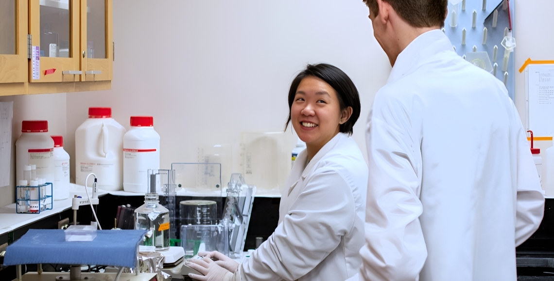 Two students in white lab coats smiling at each other while they work with test tubes