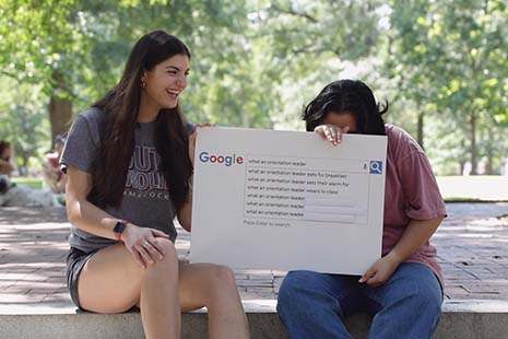 two students holding up a google autocomplete graphic, sitting outside and laughing