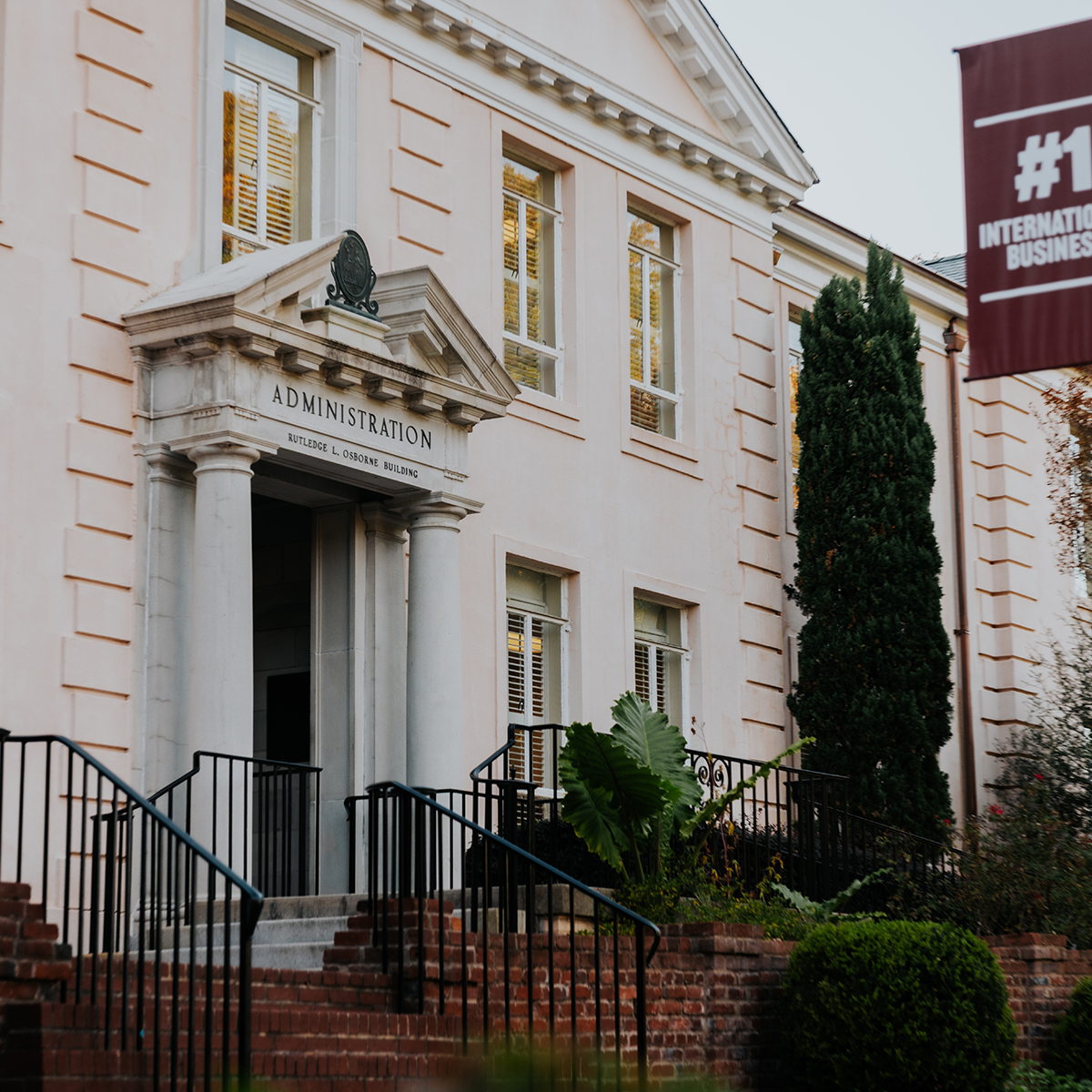 White administration building with columned entrance, brick steps, and a '#1 International Business' banner.