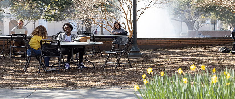 Two students hangout at a table outside of the Russell House, the Thomas Cooper fountain can be seen in the background.