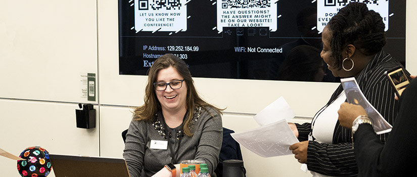Two women are laughing and looking at a computer with tech walls in the background. 