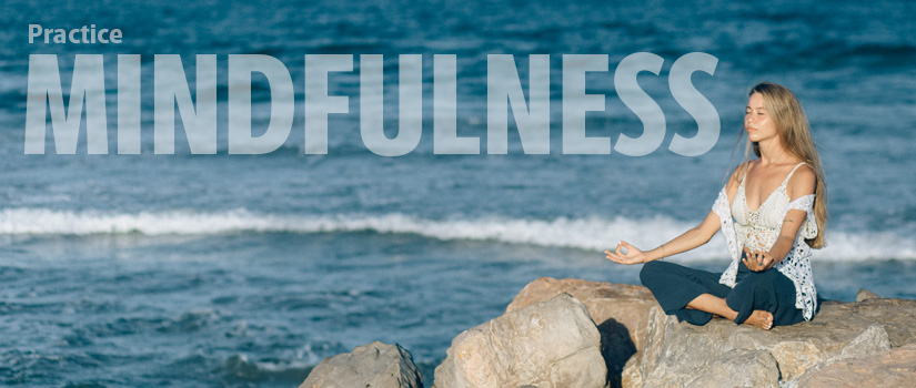 Practice Mindfulness. Woman meditates on rocks beside the ocean.