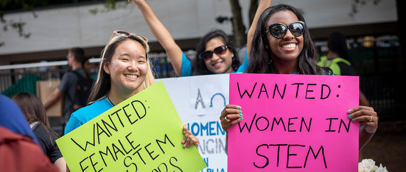 at the Greene Street Student Organization Fair, female students hold signs saying "Wanted: Female STEM Majors" and "Women in STEM"