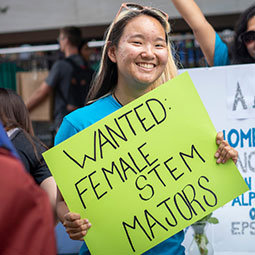 at the Student Organization fair on Greene Street, a female student holds a sign that reads "Wanted: Female STEM Majors"