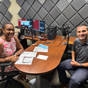 Woman and man sit inside a podcast studio with papers on the table and a red computer screen in the background.