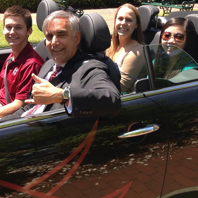 President Pastides site in the driver's seat of a convertible with three students. He is giving the "thumbs up" signal.