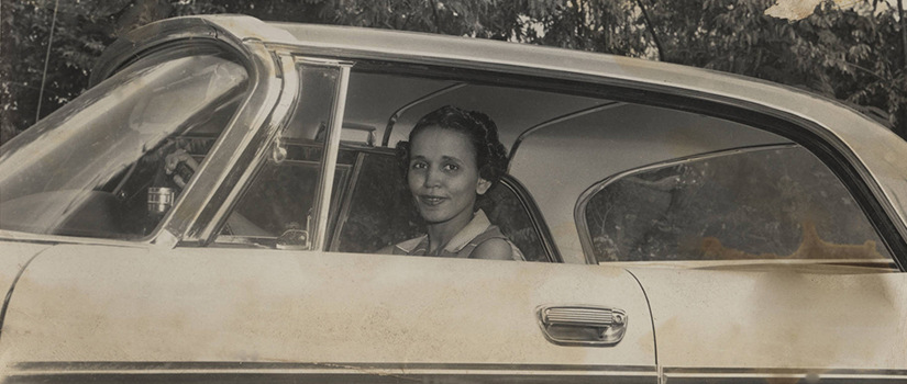 Black and white image from the 1950s or 1960s of a woman smiling in the driver's seat of a car