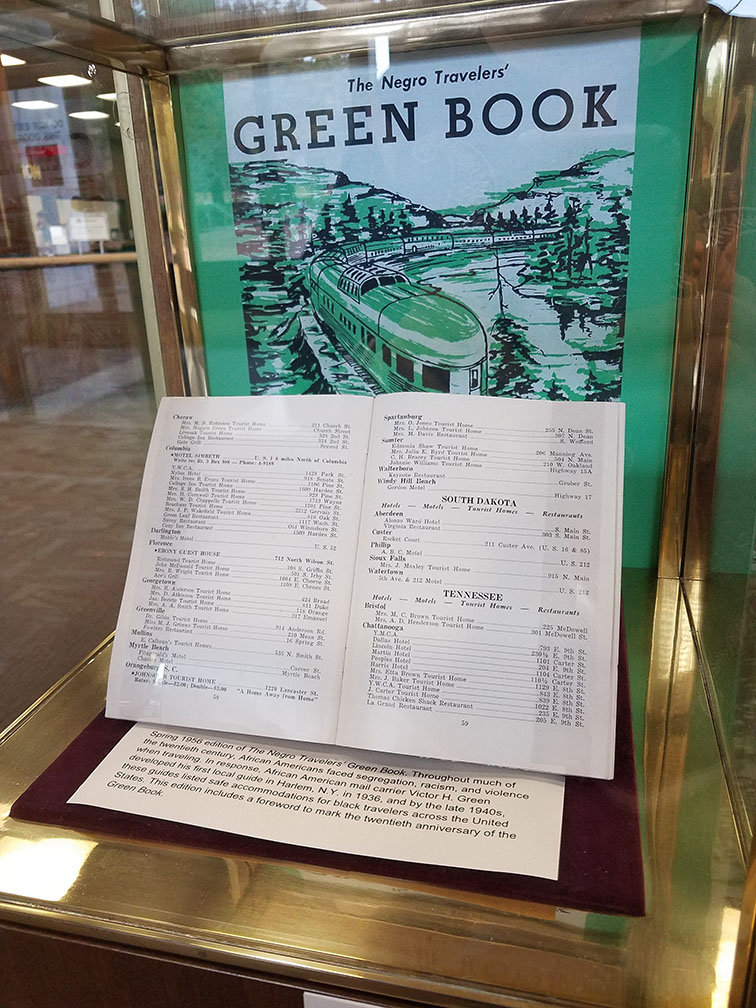 Glass display case at Thomas Cooper Library with a poster for and copy of The Negro Traveller's Green Book, the poster features a train, and the book is open to a text spread