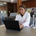 A smiling, seated person working on a laptop. The slightly angled, head-on view shows the top of the laptop, but not the screen.