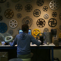 View from behind of a partially silhouetted person using film rewinds to inspect a roll of film at an inspection bench. The edges of a computer monitor facing the figure are partly visible. Empty film reels of various sizes and colors hang on the wall behind the monitor.