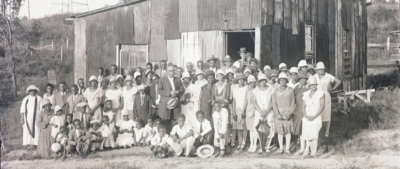 black and white image of a large group of African Americans standing for photo in front of rural building.