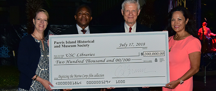 Four people outside at Beautort, South Carolina holding an oversized check for 200,000 dollars to the University of South Carolina for the preservation of the Marine Corps Film Collection from the Parris Island Historical and Museum Society