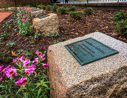 Stone markers in a flower bed surrounded by pink flowers and mulch outside of the War Memorial Building.