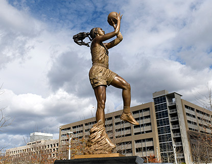 Bronze statue of A'Ja Wilson shooting a basketball with a blue sky in the backgroud. 