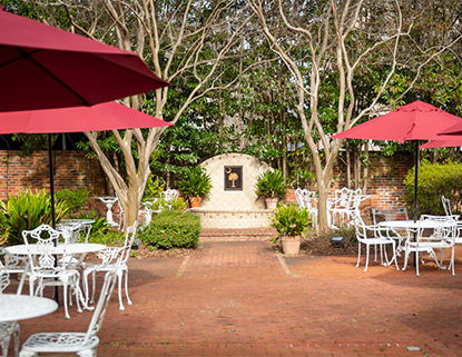 Brick patio with white tables and chairs and red umbrellas and a fountain in the back with a plaque with a palmetto tree on it. 