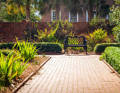 Brick walkway with a wrought iron bench at the end surrounded by flowering beds. 