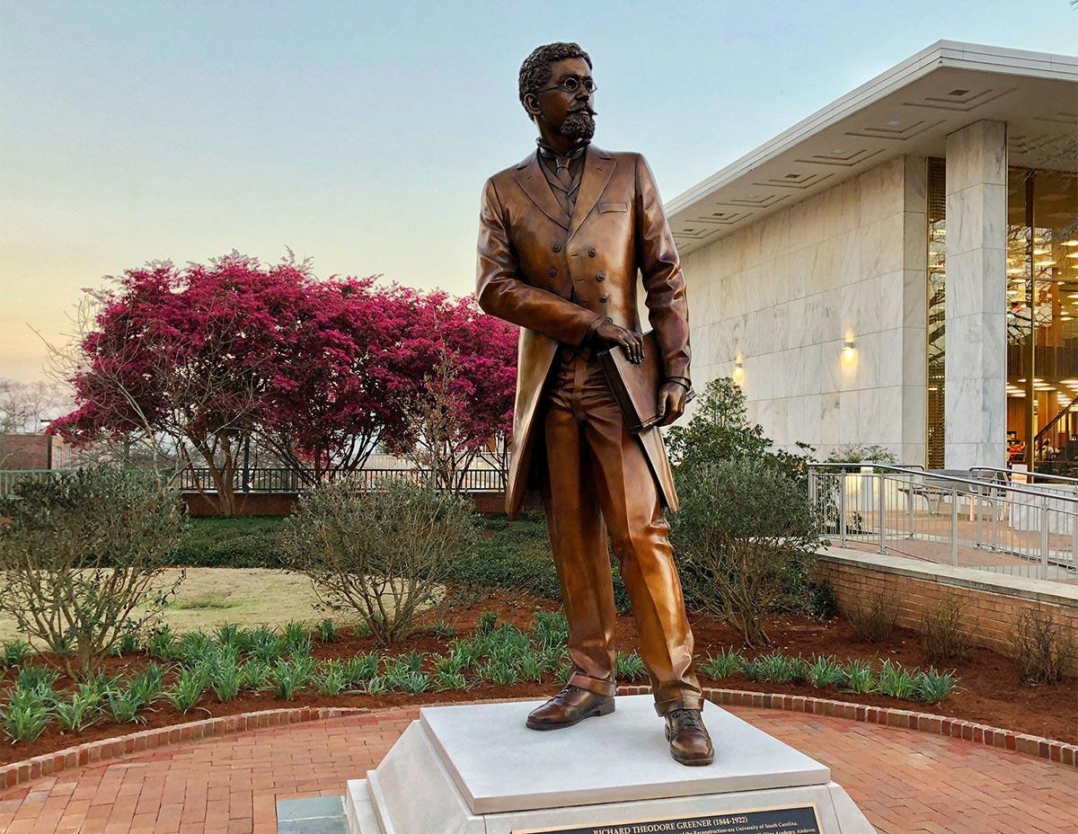 Statue of Richard T. Greener, the first African-American professor at the University of South Carolina, with pink flowers blooming and a sunset in the background. 
