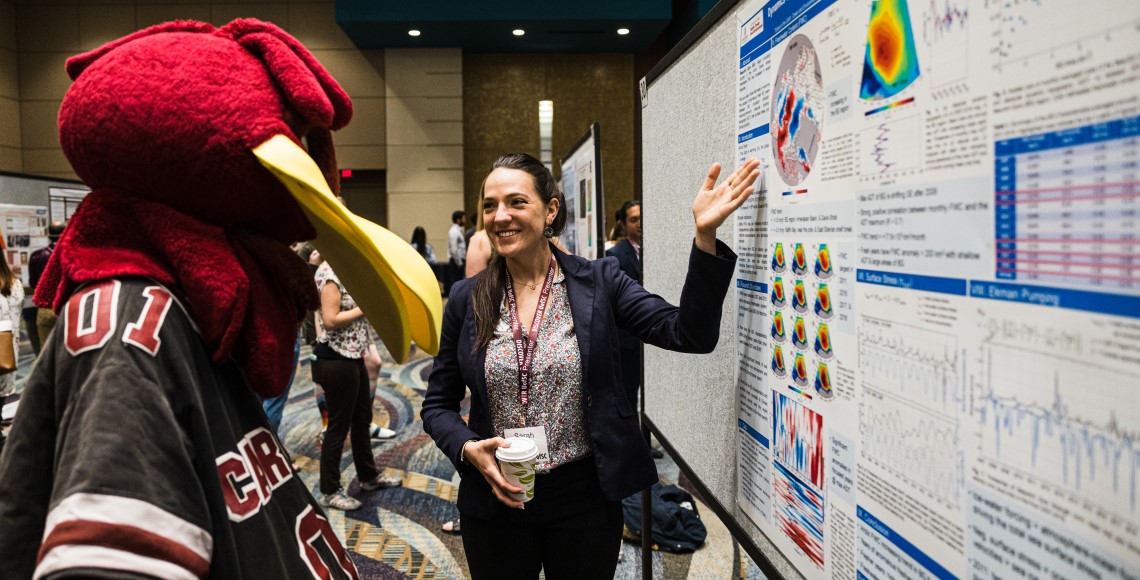 A graduate student shows her research poster to Cocky at Discover USC