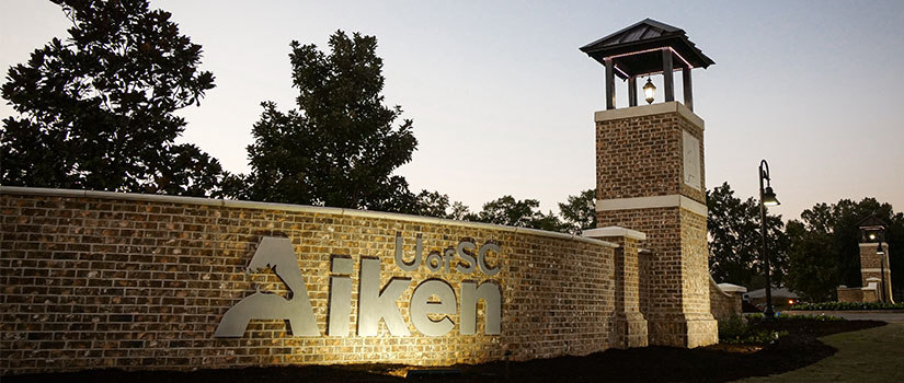 Brick entrance wall with USC Aiken sign that is lit at night