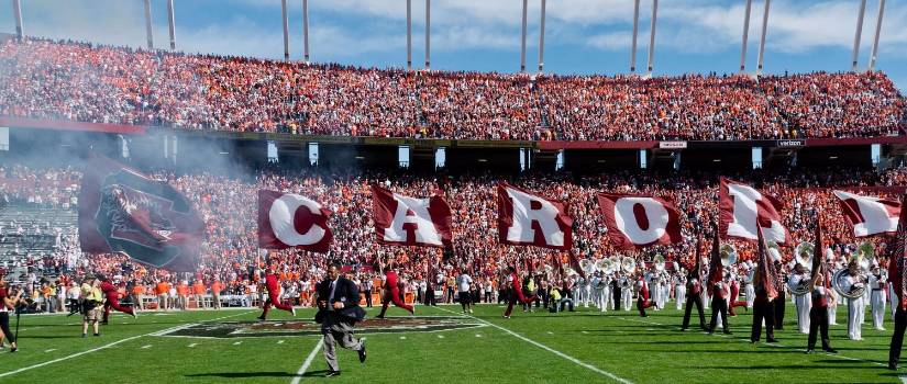 Cheerleaders running out onto football field at Williams-Brice Stadium carrying flags that show a large "Block C" logo and flags that spell out Carolina, letter by letter, with the band and fans in the background
