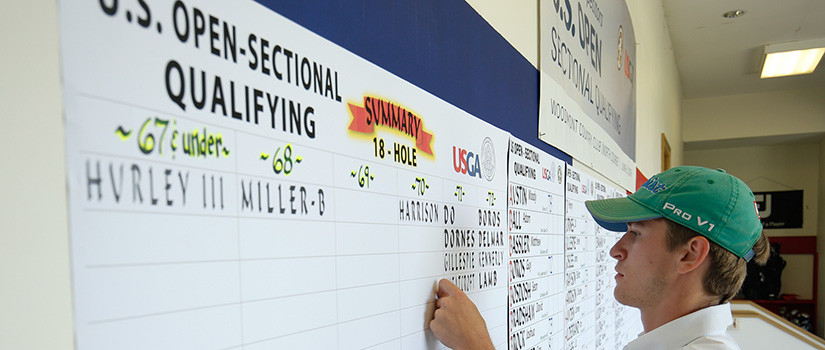 Student intern, wearing a green baseball cap, works at a U.S. Open qualifying golf tournament, posting score updates on a wall 
