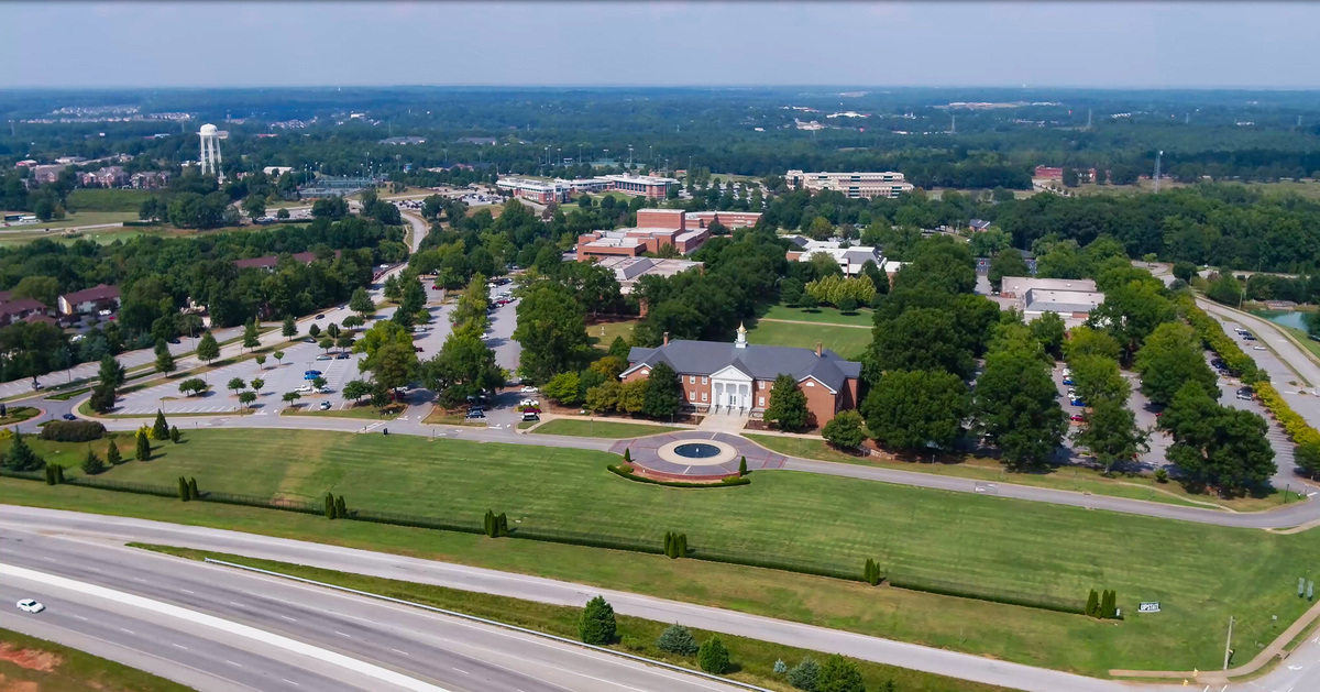 picture shows an aerial view of the upstate campus. Several brick buildings surrounded by trees are shown. At the top of the picture is a skyline view with several buildings in the distance and a water tower.
