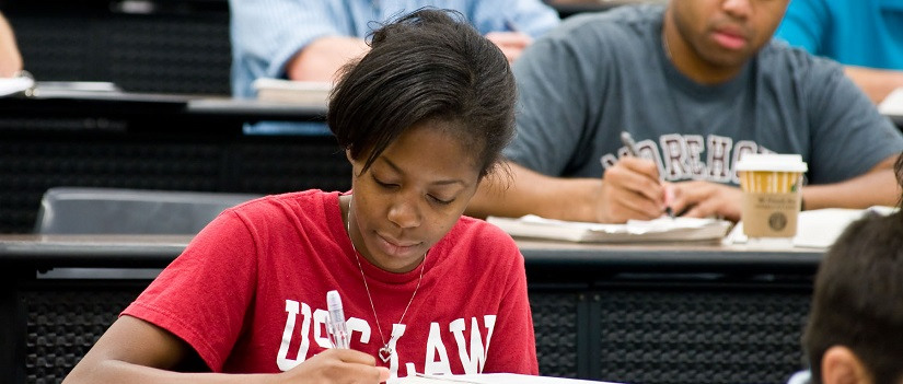Female student wearing a red T-shirt that reads USC Law takes notes at a desk in a law school classroom